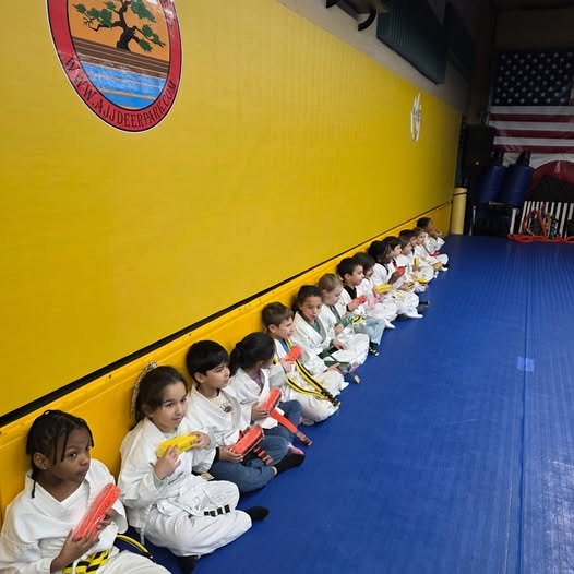 Kids lined up along the yellow wall in white uniforms before class at Makoto Jiu Jitsu Centers in Deer Park
