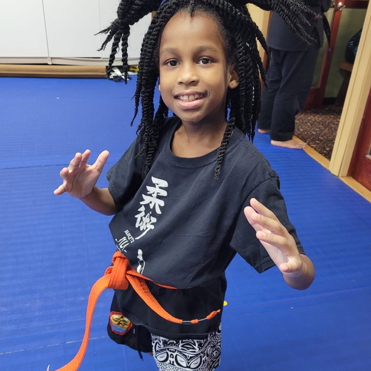 A smiling student in a black uniform and orange belt on the blue mats at Makoto Jiu Jitsu Centers in Deer Park